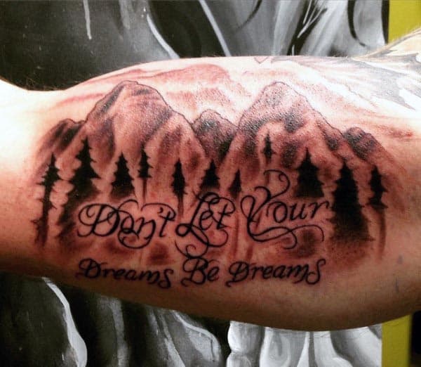 Black and grey tattoo of mountains and pine trees with cursive script reading 'Don't Let Your Dreams Be Dreams' on the inner bicep