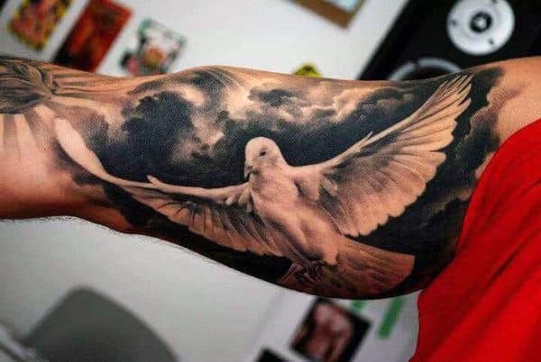 Tattoo of a detailed dove with spread wings on a bicep, with a cloudy background