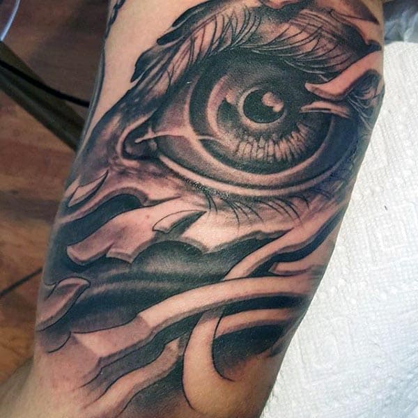 Black and grey tattoo of a highly detailed eye with surreal shading and abstract elements blending into the design on the upper arm