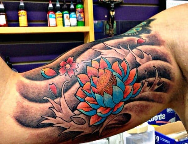 Vibrant lotus flower tattoo with blue and orange petals, surrounded by flowing leaves and floral accents on the inner bicep