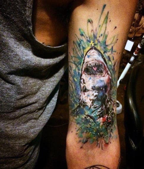Colorful watercolor-style tattoo of a shark with splashes of blue, green, and yellow, featuring a heart detail inside its mouth