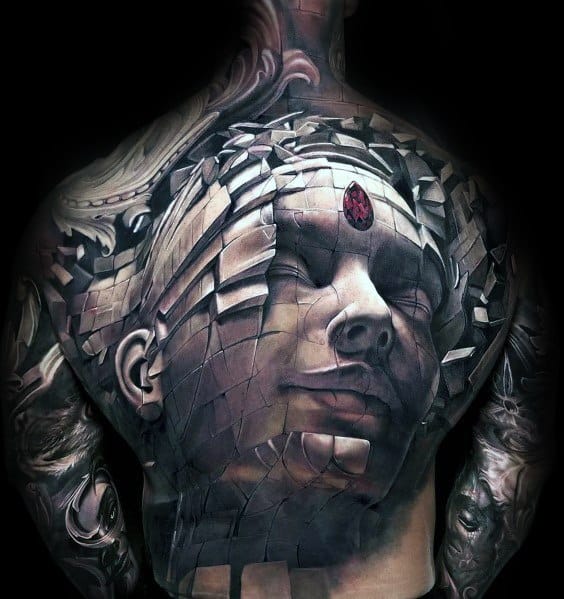 Large back tattoo featuring a 3D fragmented stone face with a red gemstone on the forehead, creating a surreal and sculptural effect.