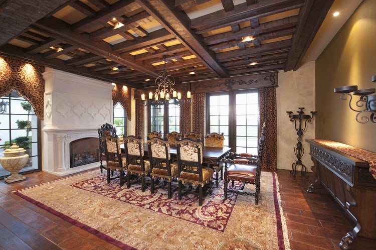 Elegant dining room with ornate wooden ceiling, large table, upholstered chairs, intricate rug, and cozy fireplace