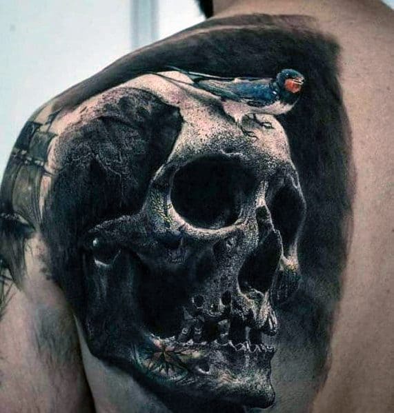 Realistic shoulder tattoo featuring a detailed skull with a perched swallow, incorporating dark shading and subtle color for a striking contrast.