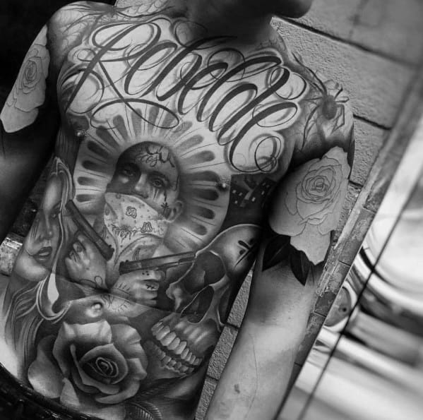 Black and grey full-torso tattoo featuring the word "Rebelde," a masked figure with a halo, guns, skulls, roses, and detailed shading for a bold design.