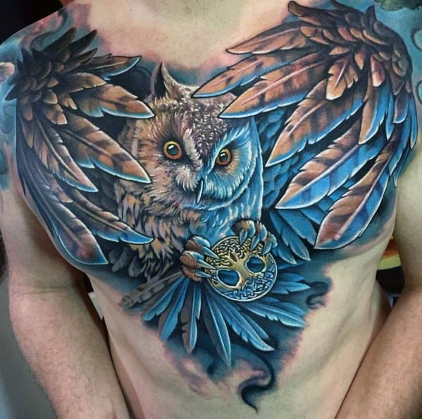 Vibrant full-chest tattoo featuring a detailed owl with outstretched wings, piercing eyes, and a decorative mask in its talons, using rich blue tones.