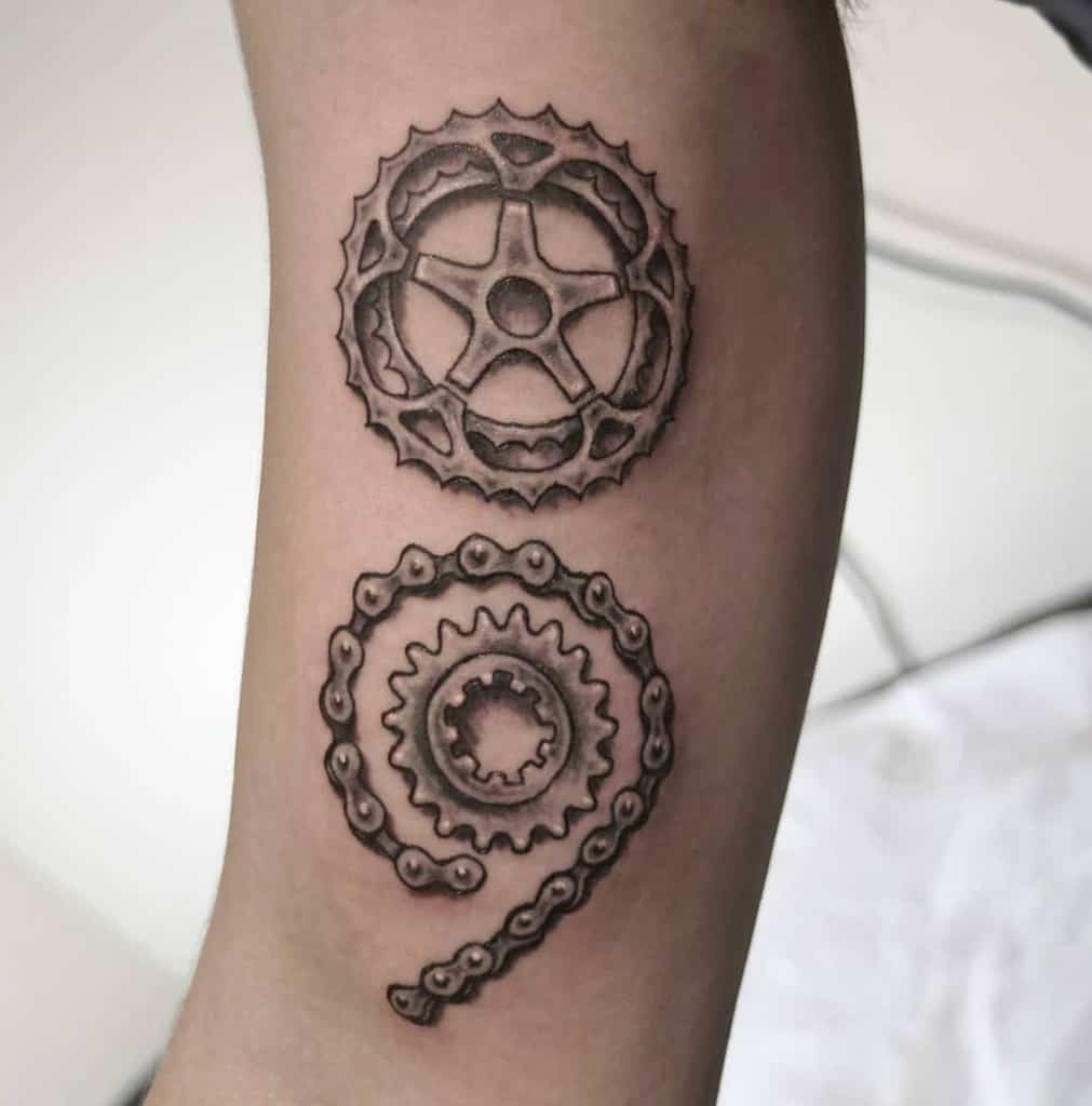 The tattoo features a detailed design with a gear at the top and a chain forming the shape of a semicolon. The mechanical elements create a strong, intricate appearance.