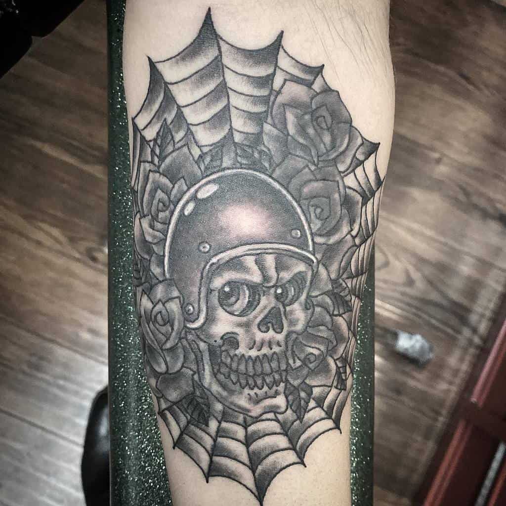 Black and grey tattoo of a skull wearing a helmet, surrounded by roses and a spiderweb, blending biker and gothic styles on the leg.