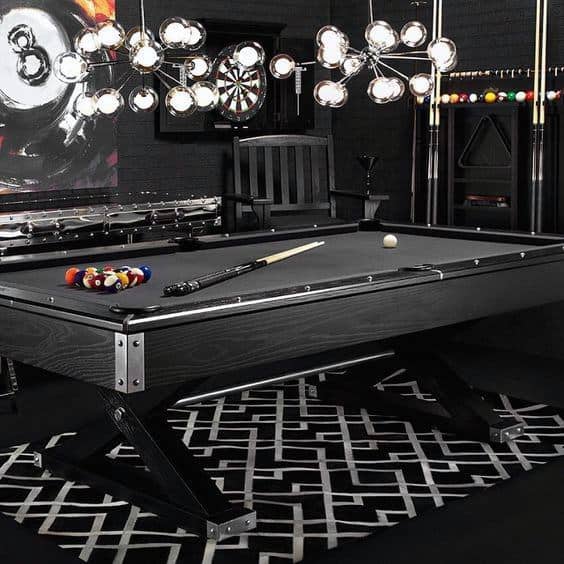 Dark masculine billiard room with black pool table, geometric rug, and modern chandelier lighting.