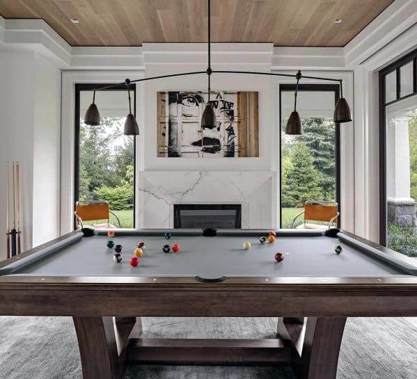 Modern billiard room with gray felt pool table, marble fireplace, and large windows overlooking greenery.