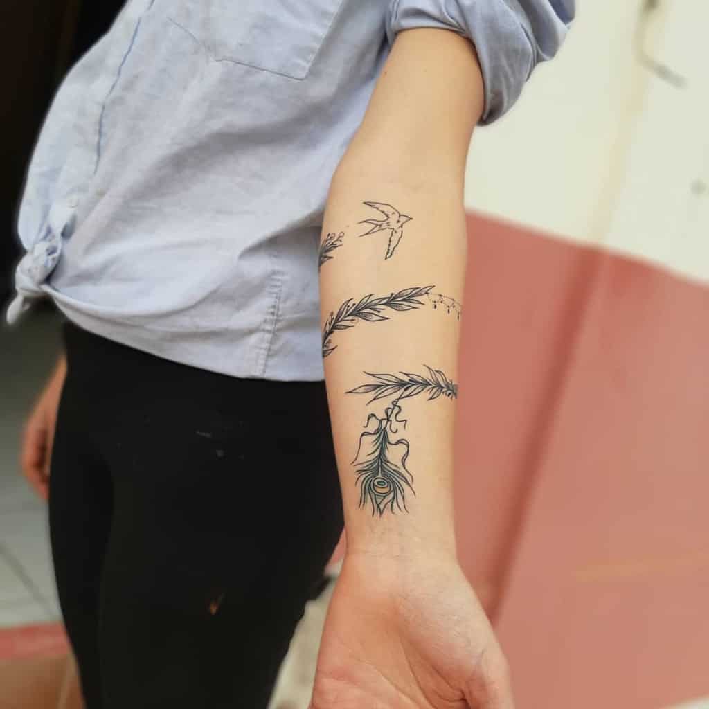 Minimalist forearm tattoo featuring a peacock feather, delicate branches, and a small bird, created with fine black linework and subtle details.