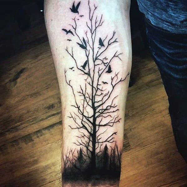 Forearm tattoo of a leafless tree with birds flying against a dark background