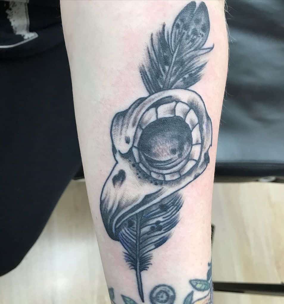 Black and gray tattoo of a skull with an exposed eye socket and a feather, placed on the forearm.