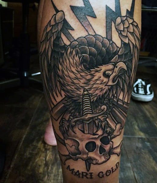 Tattoo of an eagle clutching a dagger-pierced skull with "Mari Gold" text on leg
