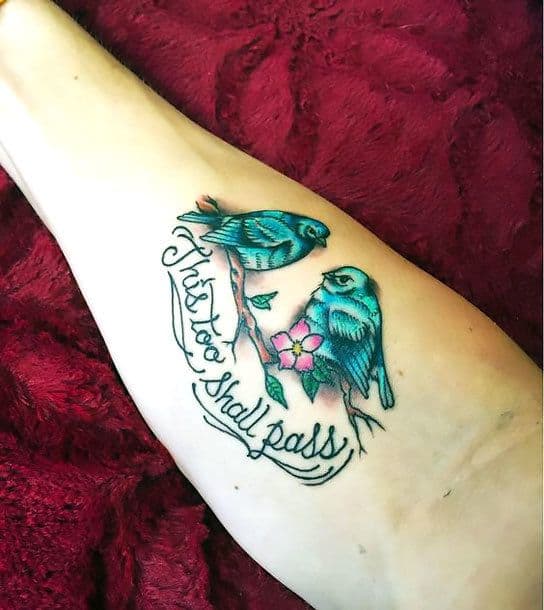 This tattoo features the phrase "This too shall pass" in flowing script, accompanied by two bluebirds perched on a branch with pink flowers. The design combines bright colors and soft details, adding a touch of nature to the inspirational quote.