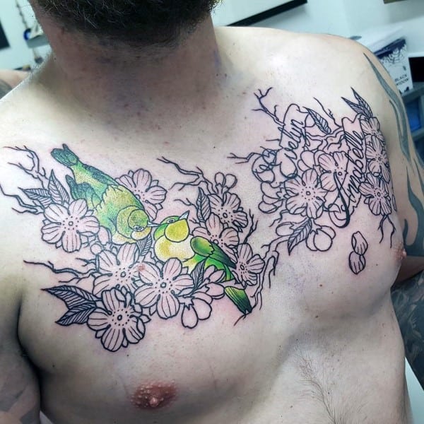 Chest tattoo featuring two vibrant green birds perched on cherry blossom branches, with delicate pink flowers and flowing script detail