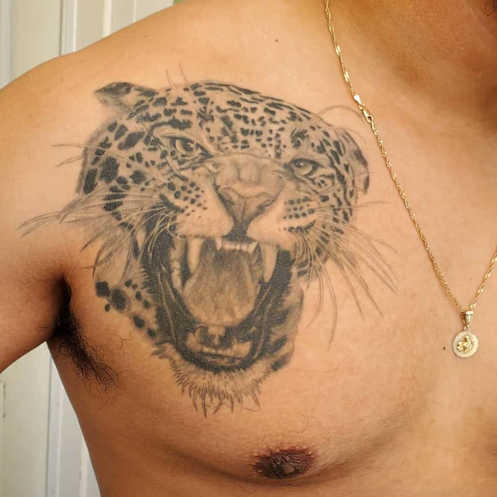 A fierce black and grey tattoo of a jaguar's roaring face, covering the chest and shoulder with detailed fur and facial features.