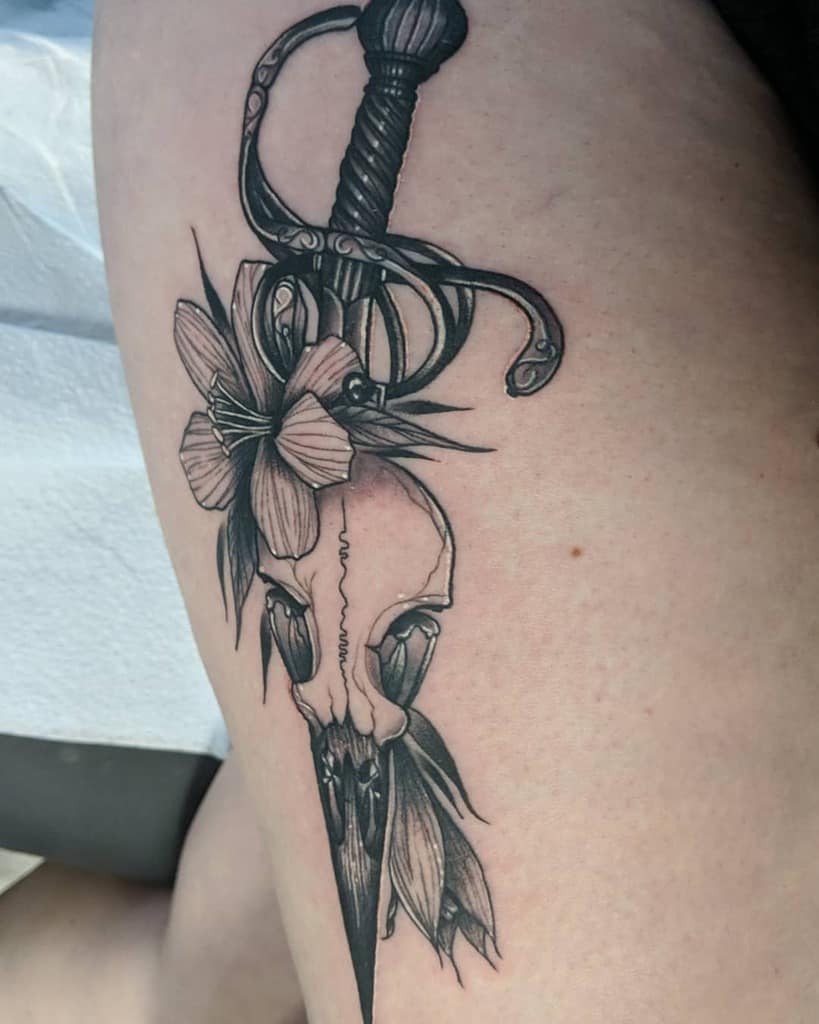 Detailed black and grey tattoo of a rapier piercing an animal skull, adorned with lilies, symbolizing strength, mortality, and elegance