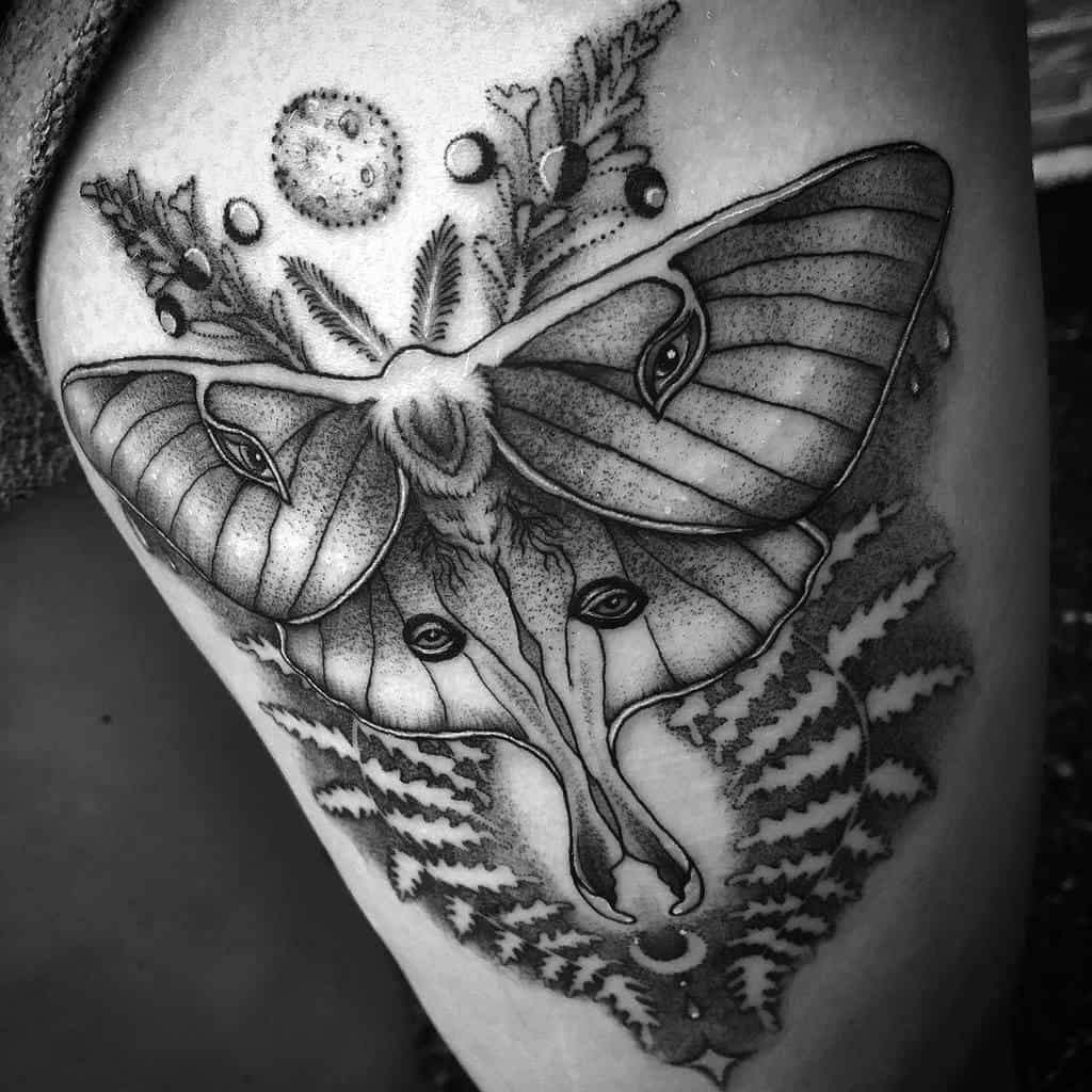 A black and grey tattoo featuring a Luna Moth with multiple eyes on its wings, surrounded by moon phases and ferns