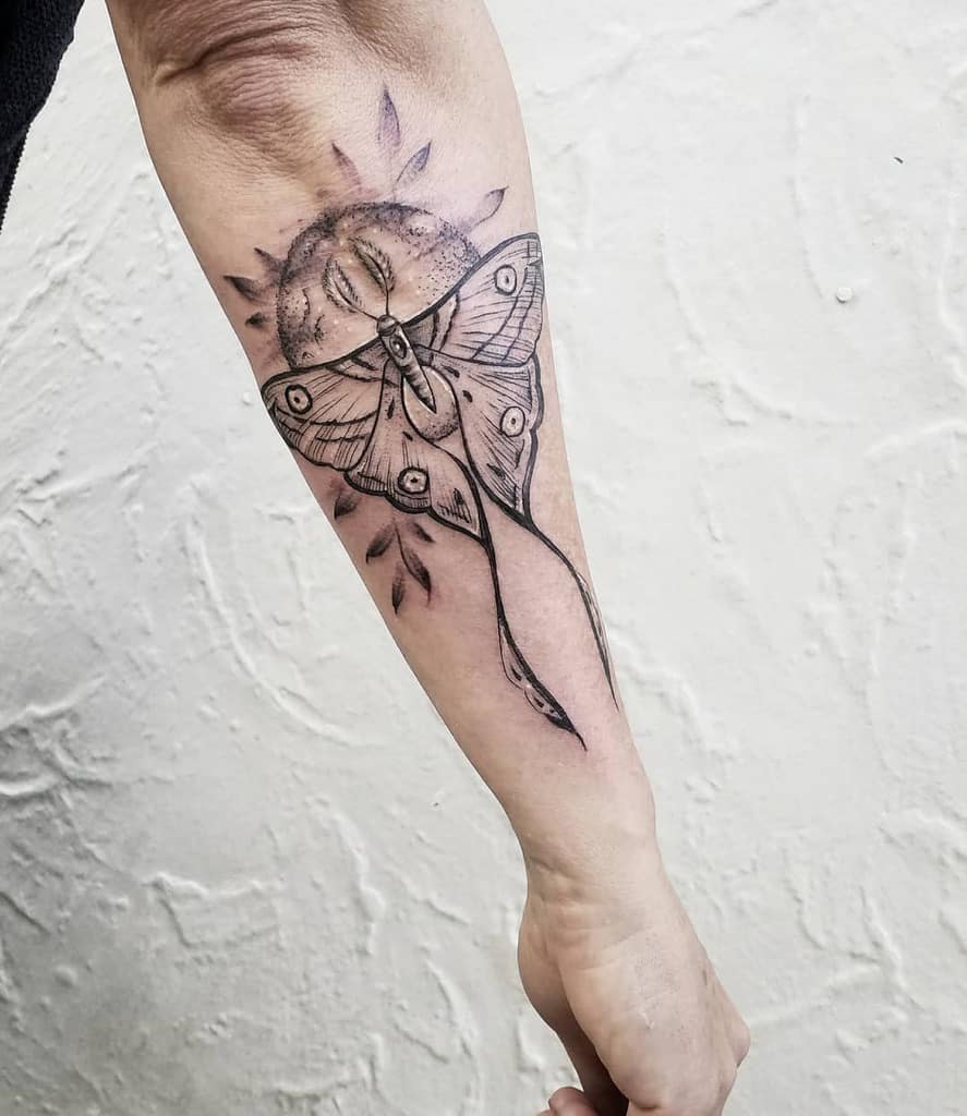 A black and grey tattoo featuring a Luna Moth in front of a circular sun design, with intricate detailing on the wings and sun rays