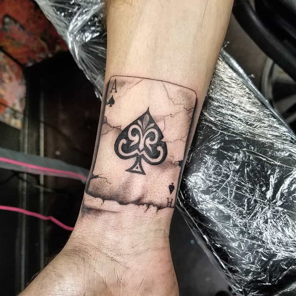 A black-and-white tattoo of an Ace of Spades card with intricate detailing inside the spade, surrounded by cracked effects