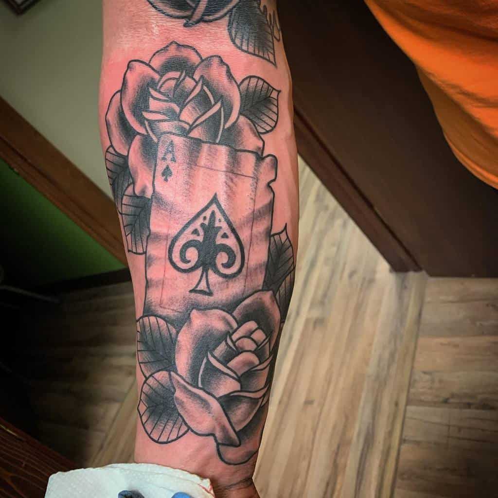 A black-and-white tattoo of the Ace of Spades card surrounded by detailed roses and leaves