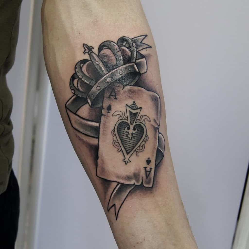 A black-and-white tattoo featuring the Ace of Spades card with a heart design, crowned with a detailed crown and flowing ribbon