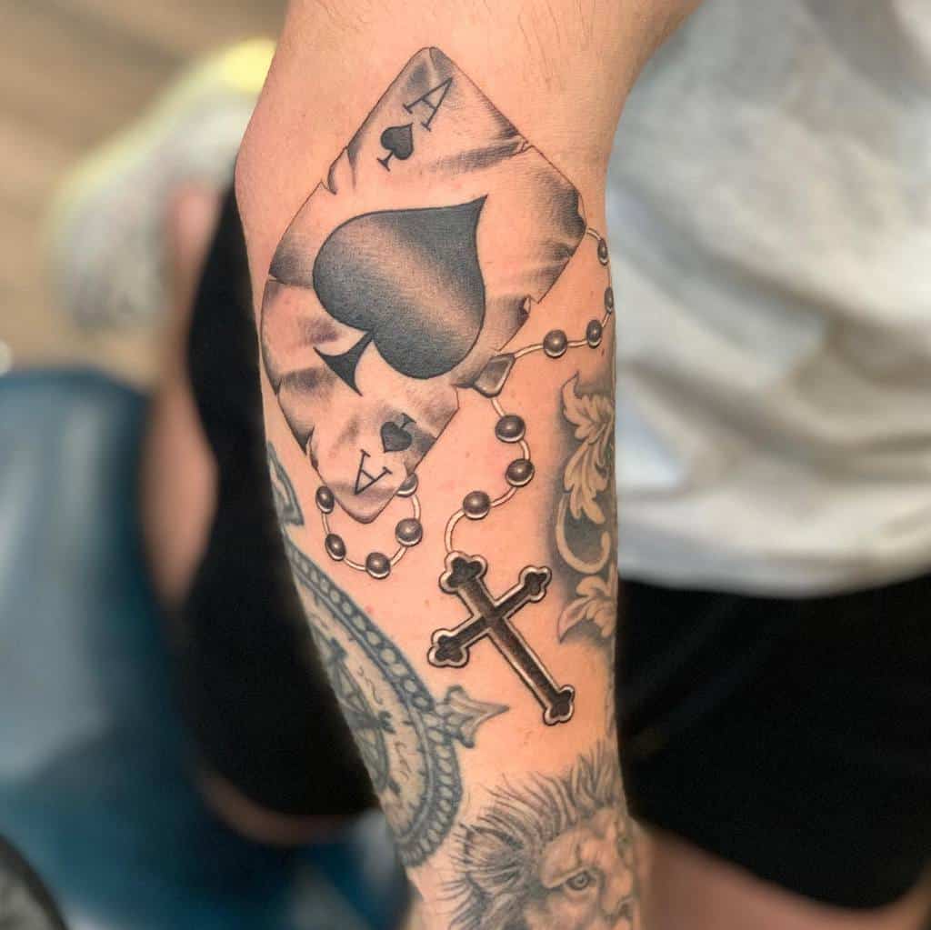 A black-and-white tattoo of the Ace of Spades card surrounded by a rosary with a cross