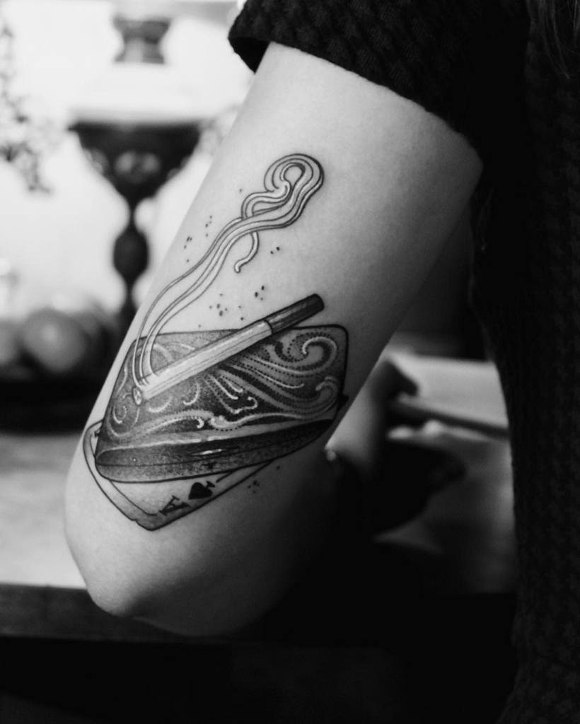 A detailed black-and-white tattoo of a waves design suitcase with cigarette smoke rising and an Ace of Spades symbol