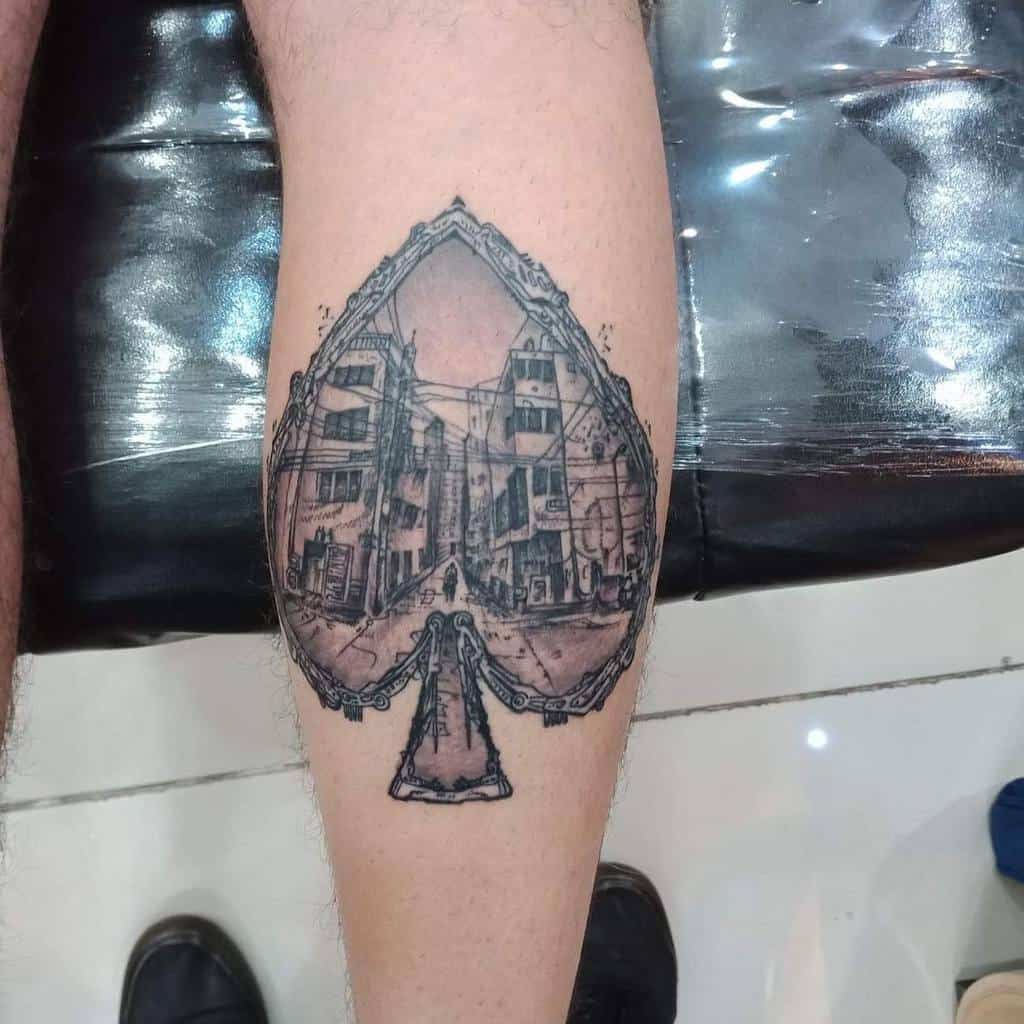 A black-and-white tattoo of an Ace of Spades card with an intricate street scene inside, framed by ornate detailing