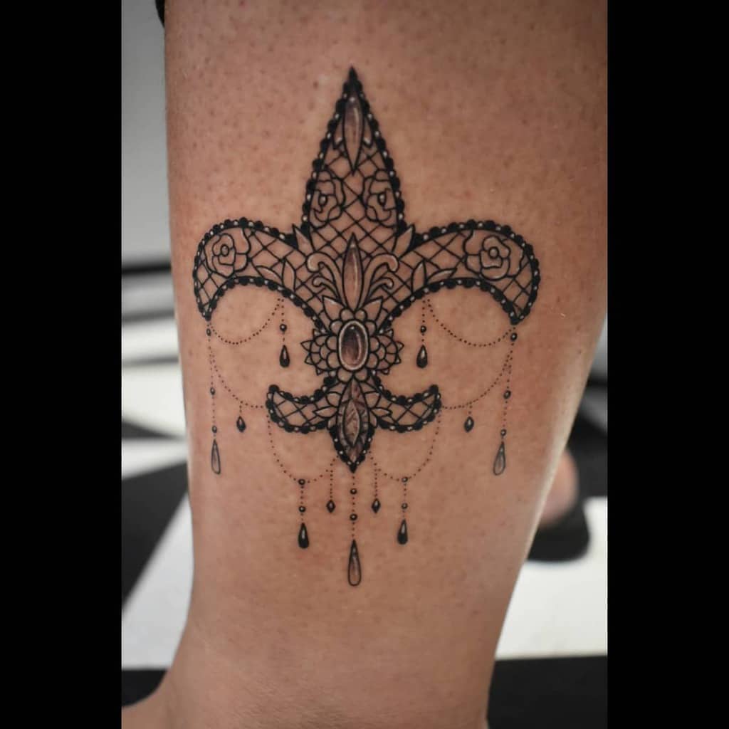 Intricate black ink fleur-de-lis tattoo with lace patterns, ornamental details, and delicate beaded chain accents.