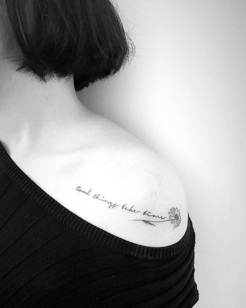 Shoulder tattoo small black and grey cursive script ‘Good things take time’ leading to fine line daisy