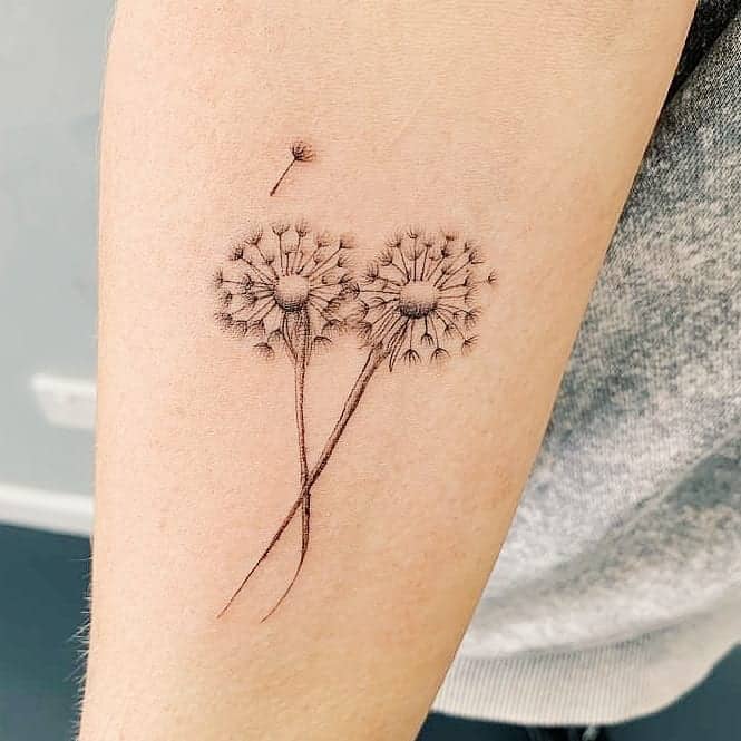 Minimalist dandelion tattoo with fine-line detailing, featuring two dandelions with seeds blowing away, symbolizing freedom and wishes.