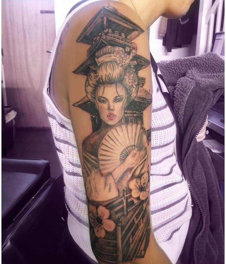 Intricate Japanese-style arm tattoo of a Geisha holding a fan, with pagoda structures woven into her elaborate hairstyle, accented by cherry blossoms.