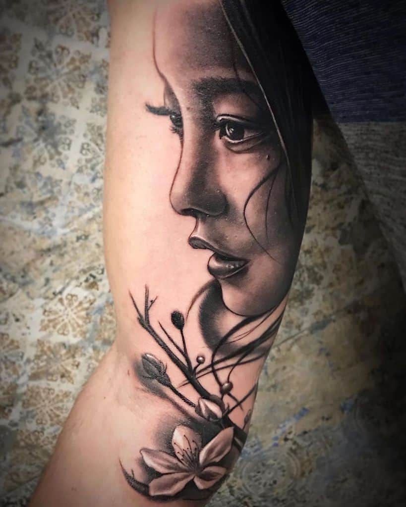 Realistic black and gray tattoo of a woman's face in profile, with flowing hair and delicate cherry blossoms blending into the design.