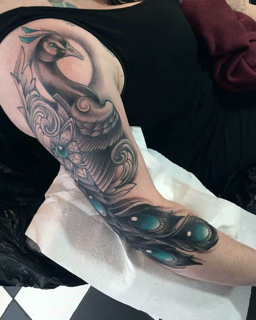 Detailed peacock tattoo with a flowing tail and teal accents, blending realistic shading and soft curves on the arm.