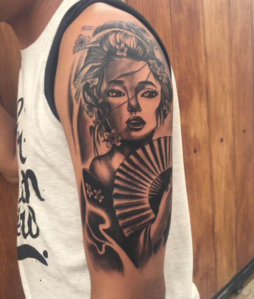 Black and gray upper arm tattoo of a Geisha with intricate shading, floral hair ornaments, and a detailed fan, blending realism and traditional Japanese art.