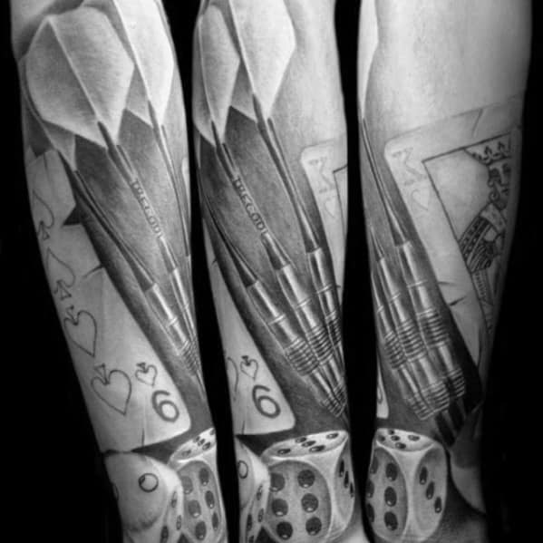 Realistic forearm tattoo with detailed darts, dice, playing cards, and a six of hearts, blending themes of skill and chance.