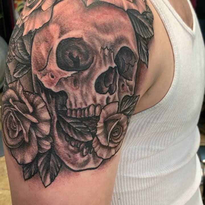 Black and grey shoulder tattoo of a skull surrounded by detailed roses, featuring bold shading and intricate linework for a dramatic look.