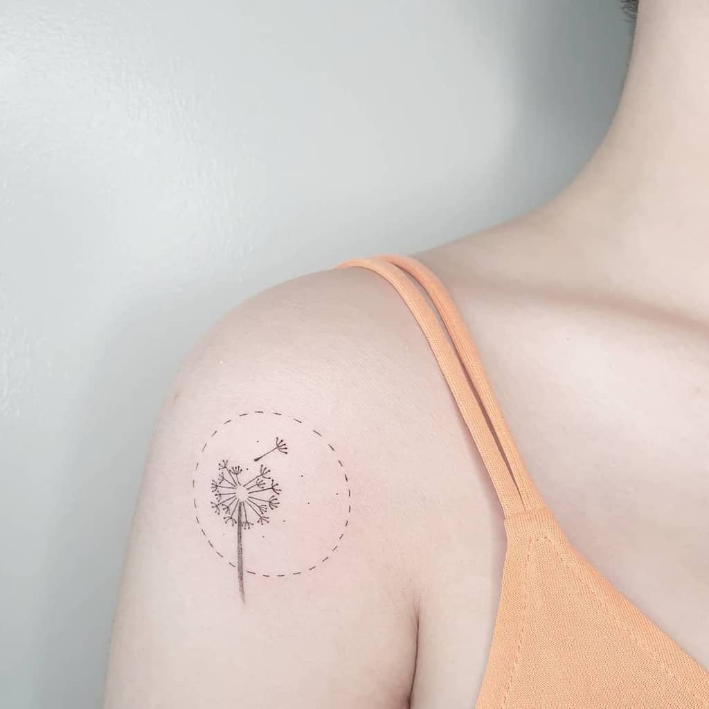 This tattoo features a delicate dandelion with seeds drifting away, enclosed within a soft dotted circle. The design evokes lightness and freedom, emphasizing the beauty of movement and change.