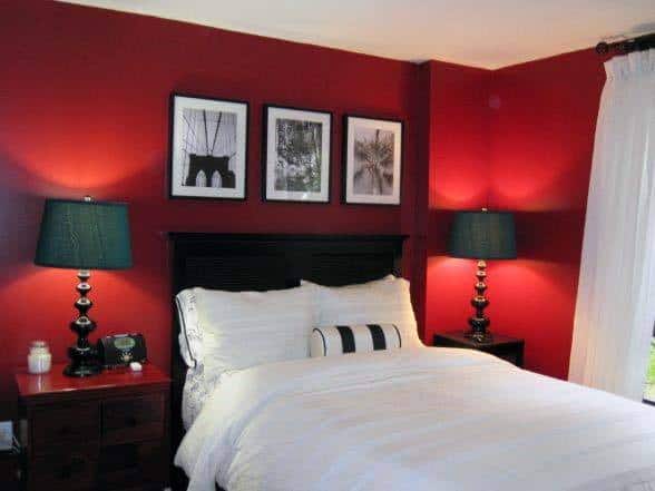 A red-walled bedroom with a white bed, black lamps, and framed monochrome photos above the headboard