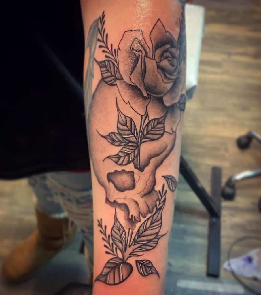 Elegant black and grey tattoo of a skull intertwined with roses and leaves, featuring fine linework and soft shading on the forearm.