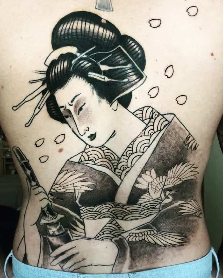 Large black and gray back tattoo of a Geisha holding a katana, wearing a patterned kimono with cranes, surrounded by falling cherry blossoms.