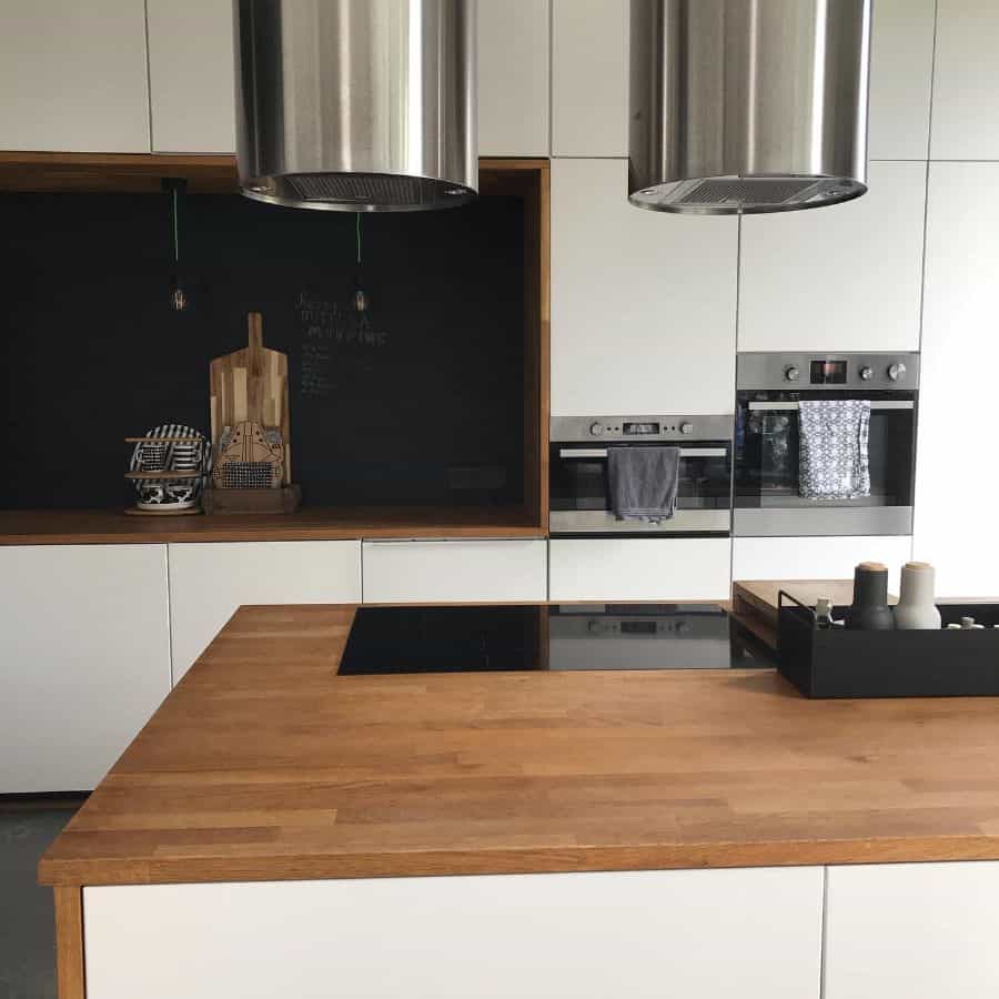 Modern kitchen with wood countertop island, white cabinets, black backsplash, and stainless hoods.