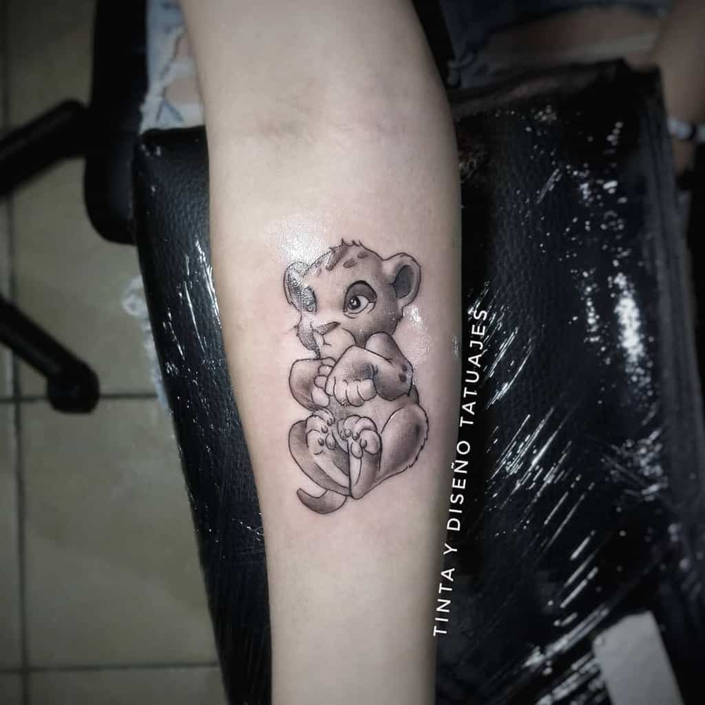 Adorable tattoo of Simba as a cub sitting comfortably, captured in black and gray shading to highlight his playful innocence