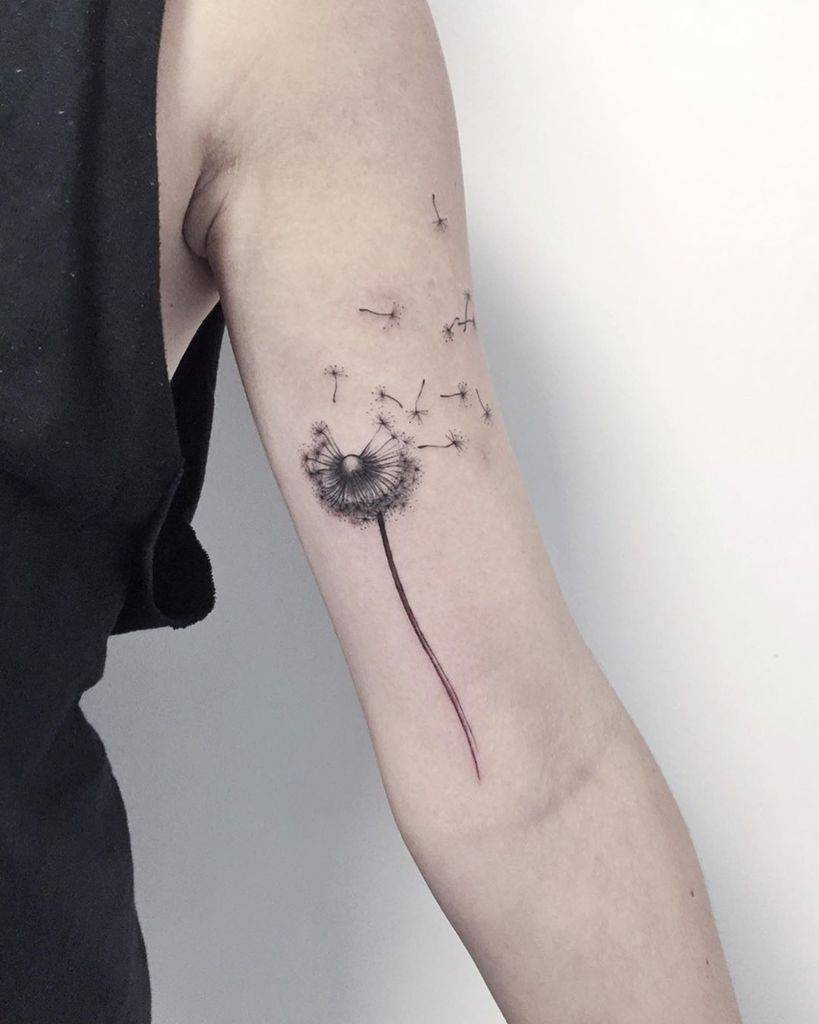 Delicate dandelion tattoo on the upper arm with seeds blowing away, symbolizing freedom, change, and letting go.