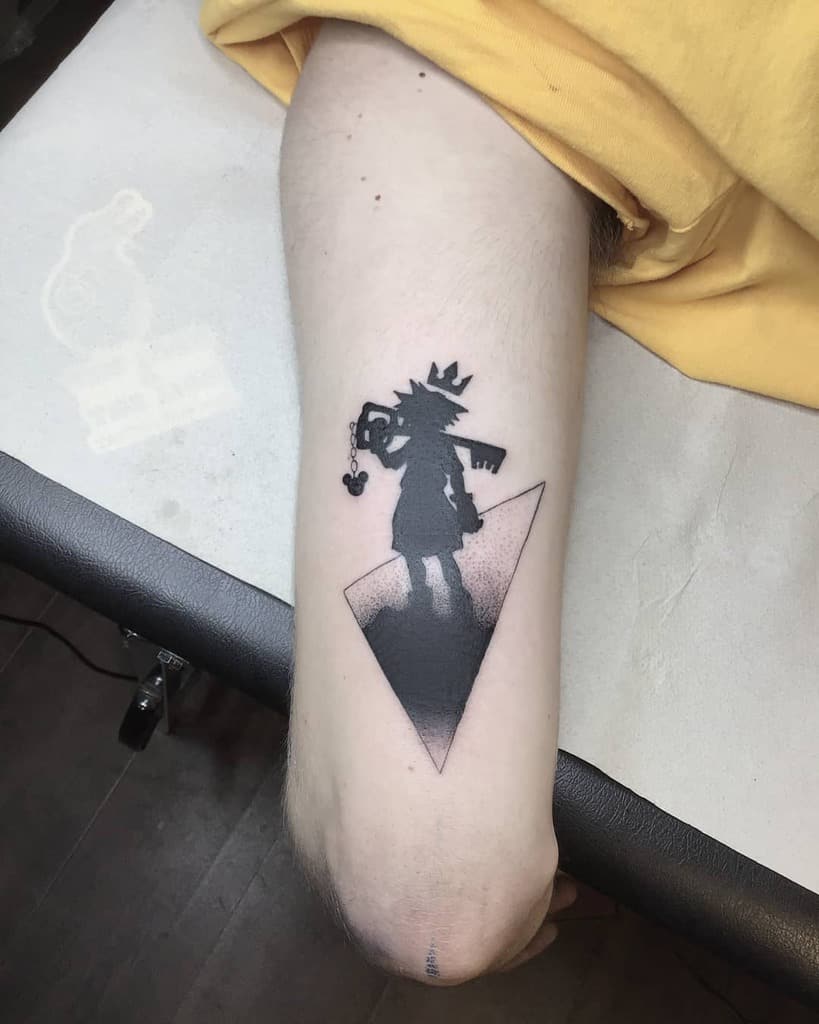 Minimalist black ink Kingdom Hearts tattoo of Sora’s silhouette holding a Keyblade, with a crown above his head and a geometric backdrop.
