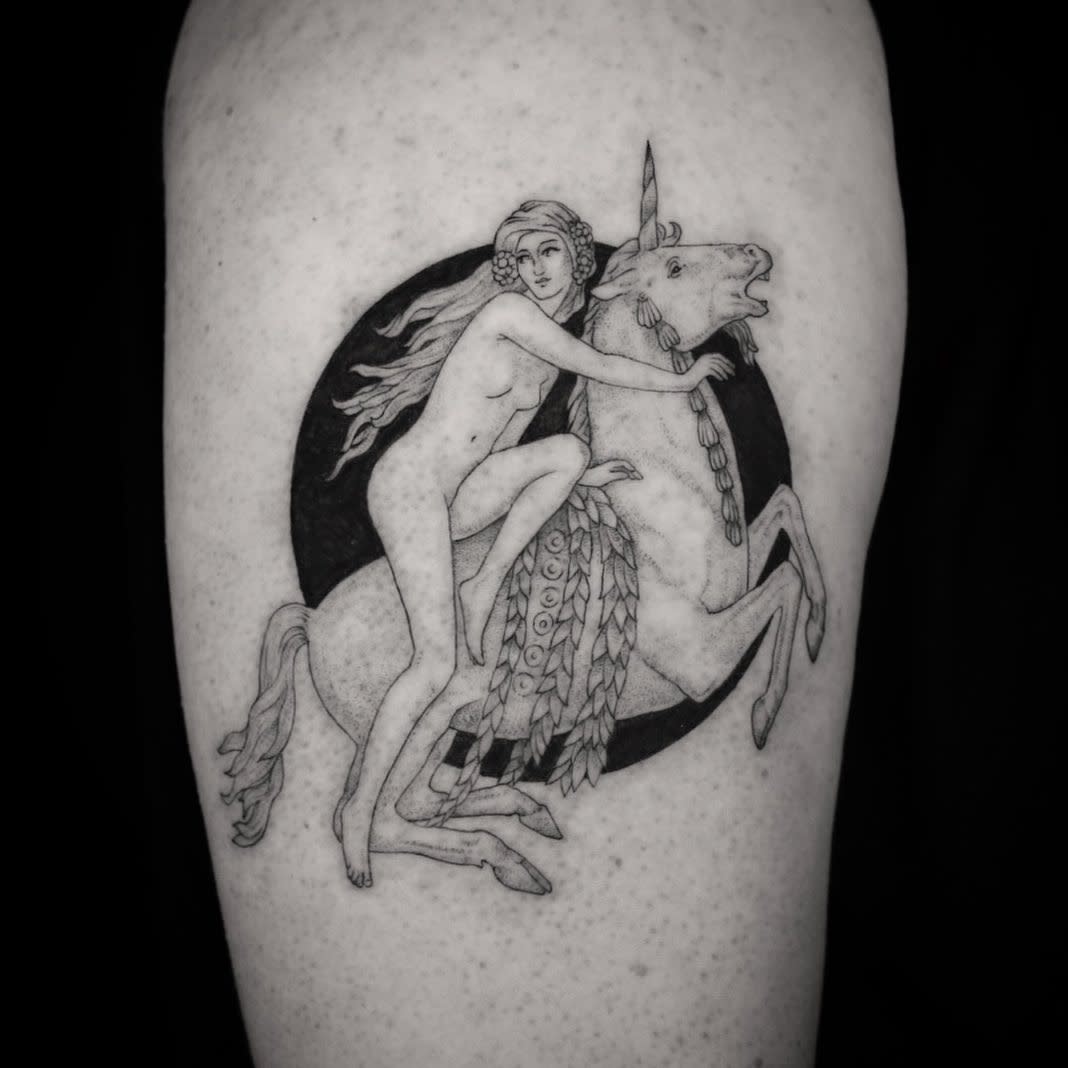 Black and grey tattoo of a mythical scene featuring a nude woman with flowing hair riding a rearing unicorn, set against a dark circular background.