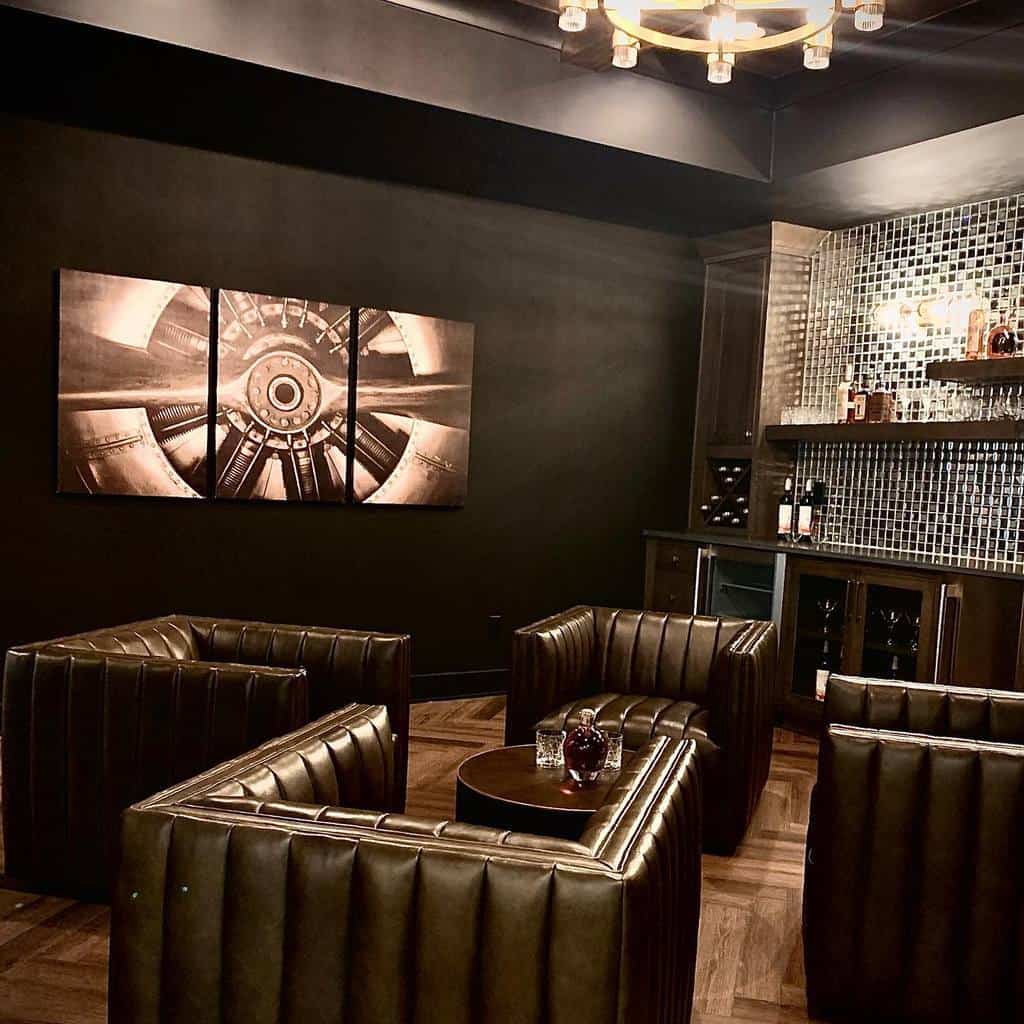 Modern lounge with black walls, leather armchairs, industrial art, and mirrored bar setup.