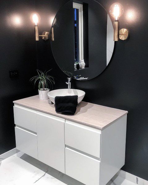 Modern black bathroom with white vanity, round mirror, gold wall sconces, and vessel sink.